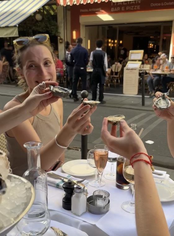A smiling guest holding up oysters at a café table with rosé Champagne