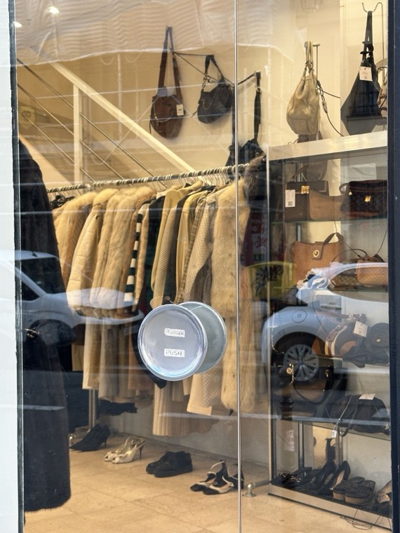 Vintage furs and coats on a rail beside shelves of handbags, seen through a boutique glass door